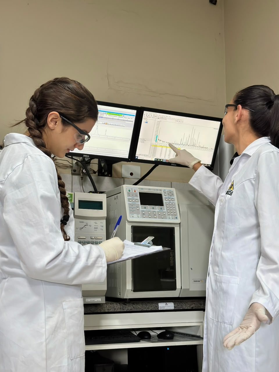 Duas analistas conferindo o cromatograma na tela, ao lado do cromatógrafo gasoso